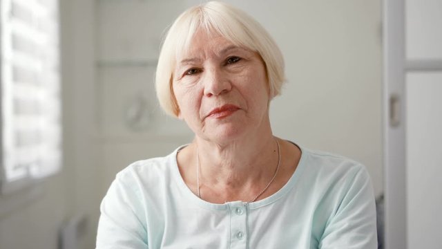 Portrait of beautiful senior blond woman at home. Sitting in white living room looking at camera smiling