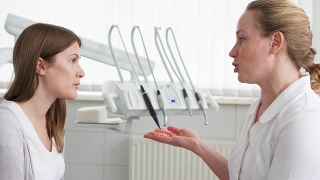 Dentist talking with woman patient in clinic. Female professional doctor at work. Showing false jaw explaining medical procedures. Dental equipment on background. Dental check up