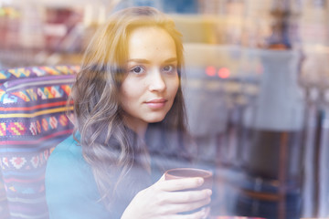 Attractive stunning young lady sitting alone at cafe table, captured through the window. Beautiful Caucasian woman relaxing on couch at modern restaurant, drinking tea, looking at camera with smile.