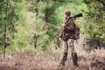 Papier Peint photo Chasser Chasseuse en tenue de camouflage prête à chasser, tenant une arme à feu et marchant dans la forêt.