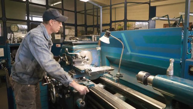Mature caucasian man working in shabby car service, making aluminum bushing on old lathe. Wearing blue jeans jacket and cap. Shot with Sony a7s and Atomos Ninja Flame indoors, dolly slide