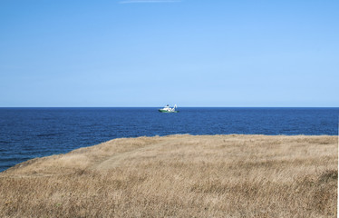 View from sea shore to calm blue sea with and fishing vessel on clear summer sky background