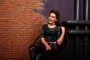 Obraz na płótnie Canvas Black girl in green shirt and leather pants with bright make-up sitting at chair on studio background brick wall. Halloween theme.