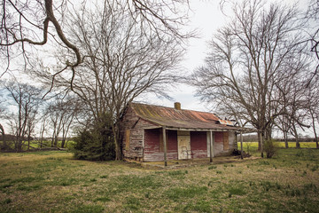 Fototapeta premium A low slung old and abandoned farmhouse with front porch veranda and it's windows boarded up and peeling paint in a rural springtime landscape