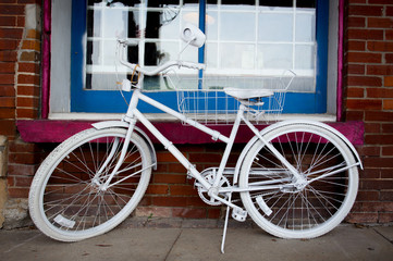Fototapeta na wymiar An old pedal bike painted white leaning on a kickstand in front of a red brick wall and multi paned window