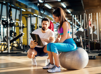 Young healthy active woman sitting on the gym ball and consulting with a  personal trainer about an...