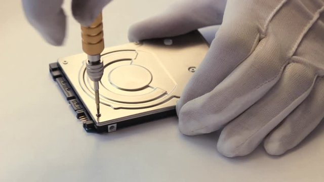 A man in white gloves twists the screw on a computer hard drive with a screwdriver.
