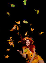 A beautiful young woman dresses up as an Autumn fairy   and wears a wreath on her head made out of Autumn leaves.  She poses for the camera while Autumn leaves fly around her.