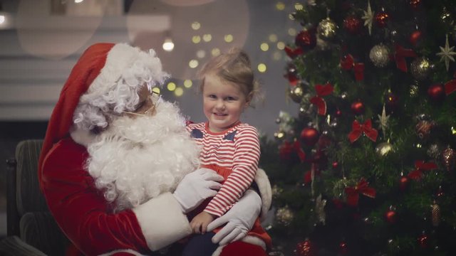 Kind Santa Claus playing with little girl sits on his knee in Christmas eve