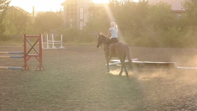 Silhouette of a young girl riding on a brown horse on a horse farm at sunset, slow motion.