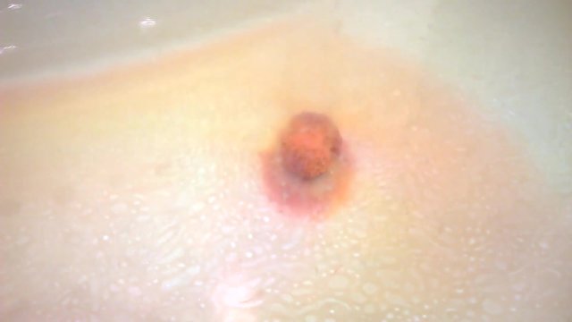 Small colorful bath bomb lies on the bottom in the tub and slowly dissolves. On the surface of the water traces of aromatic oil. The camera moves. Focus on oil