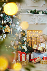 Little girl with giftbox sitting on the floor of living-room by fireplace and looking at beautiful xmas tree