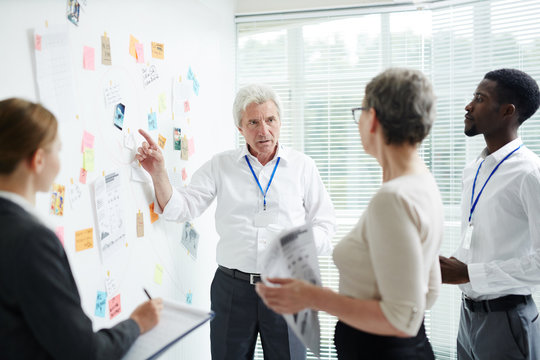 Multi-ethnic group of managers gathered together at modern boardroom and brainstorming on joint project, confident senior boss pointing at dry-erase board