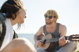 Happy guy playing guitar and singing with his friend by seaside on hot sunny day