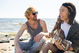 Happy guys singing by guitar on sandy beach at leisure