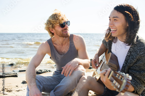 Happy guys singing by guitar on sandy beach at leisure