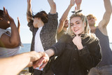 Young blond woman in black denim jacket looking at camera while following her boyfriend on background of dancing friends