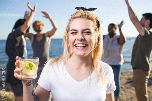 Ecstatic girl with glass of citrus cocktail enjoying gathering with friends on the beach
