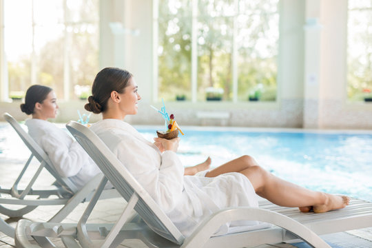 Two girls in bathrobes enjoying relax on deckchairs by swimming-pool at spa resort