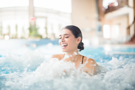 Excited female laughing while splashing warm water during spa procedure in whirlpool