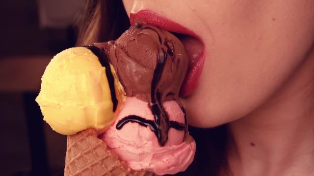 Close-up female lips with pink lipstick. Eats Italian ice cream in waffle horn with three balls yellow, pink and chocolate. Sexually bites the ice cream and licks her lips.Girl eating ice cream gelato