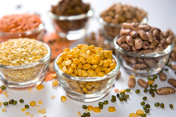 Assorted lentils and beans in glass bowls, featuring vibrant red gram (toor dal), split moong dal, and speckled rajma beans. Scattered whole legumes such as green gram and black gram add to the visual