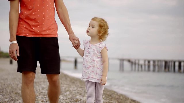 father and daughter walking hand along the sea coast. cute girl with red hair looking at your favorite parent man with a beard in a red shirt