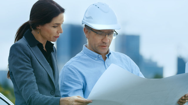 Chief Engineer Shows Project Drafts To Business Woman Investor. In the Background City with Skyscrapers.