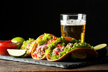 Photo of Mexican tacos with ground meat, beef, beans, onions and salsa on wooden background. Ketchup sauce and lime. A glass o beer in the background.