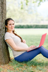 Beautiful pregnant girl with a laptop sitting under a tree. Pregnant girl looking at the camera.