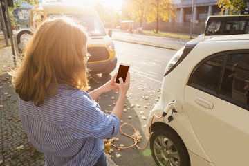 Young woman is standing near the electric car and looks at the smart phone. The rental car is...