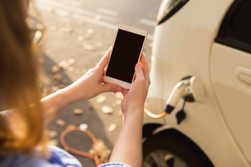 Young woman is standing near the electric car and looks at the smart phone. The rental car is...