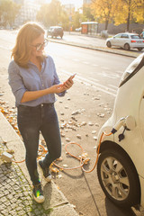 Young woman is standing near the electric car and looks at the smart phone. The rental car is...