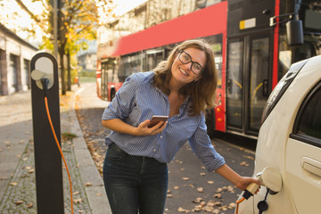 Young woman is standing near the electric car and looking at camera. The rental car is charging at...