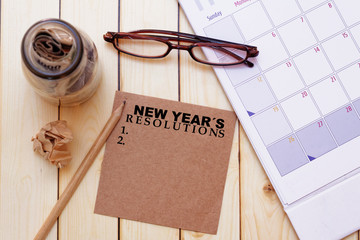 New year's resolutions concept with eye glasses,calendar, pencil,crumpled paper and a glass jar full of money on wooden background