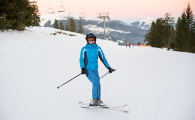 Smiling woman wearing helmet, blue sportswear and ski goggles riding skis on the snowy mountain at a winter resort with ski lifts in background.