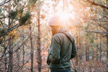 The girl is dressed in a pink hat and a green jacket stands in the woods and looks at the sunset