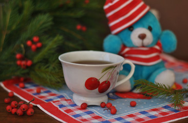 A small toy teddi bear in a red hat, spruce branches with red berries and a white cup next to these on an old wood background. Christmas and New Year cards. Still life.