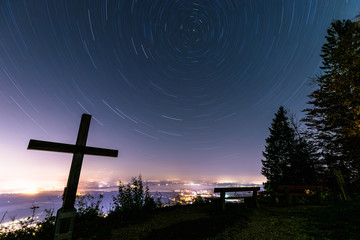 Night sky rotating over lake Chiemsee, with cross and bench, long time exposure, view from Schnappenkirche