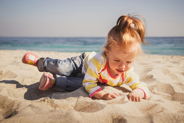 Small baby, little girl in blue jeans, pink shoes and colourful pullover sitting and playing in sand at the beach