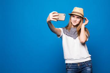 Young cheerful attractive woman on the blue background. She is taking selfie on the camera of her phone, wearing casual summer outfit and a hat