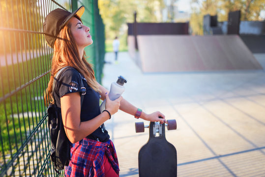 Cute urban girl with long-board in skate-park drinking water