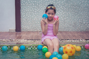 Asian little cute girl sitting on pool edge, wearing pink swimminh suit and her hand holding small ball.