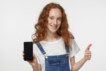 Fototapeta premium Indoor portrait of attractive redhead girl isolated on grey background, holding blank smartphone, smiling at camera, showing thubms up gesture, feeling happy