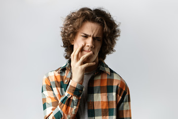Picture of stressed out young male wearing stylish plaid shirt having painful expression, frowning and touching face, suffering from toothache. People, dental care and health problems concept