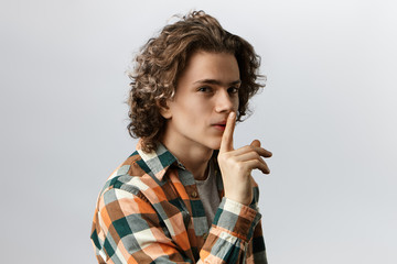 Cute stylish guy dressed in plaid shirt making silence gesture, keeping finger at his mouth and saying Shh, asking to keep silence while planning surprise birthday party for his girlfriend. Top secret