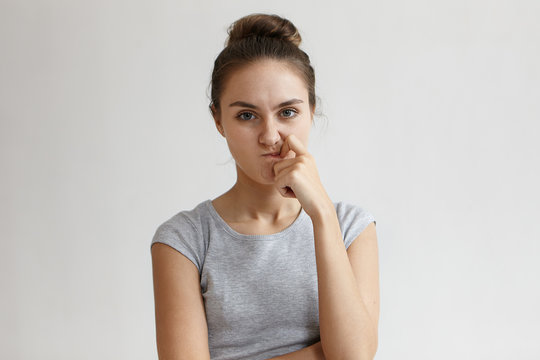 Let me think. Picture of serious attractive female student in grey t-shirt keeping finger on her lips and frowning, having pensive frustrated look while working on research for diploma project