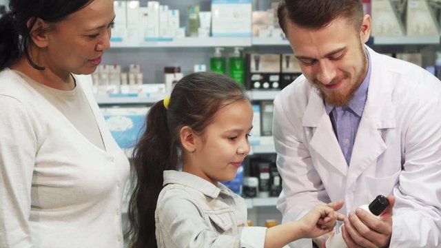 The pharmacist holds one of the tubes in his hands. A little girl stands next to him and listens to what he tells her. The chemist looks very nice and friendly. Mom and daughter are in the pharmacy