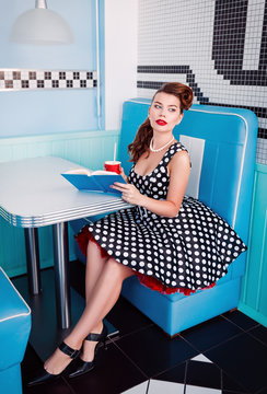 Retro (vintage) portrait of pretty young girl sitting in cafe with book and beverage. Pin up style portrait of young girl in dress