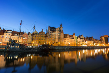 Scenic view of a tourist sailing boat and lit old buildings along the Long Bridge waterfront at the Main Town in Gdansk, Poland, in the evening.